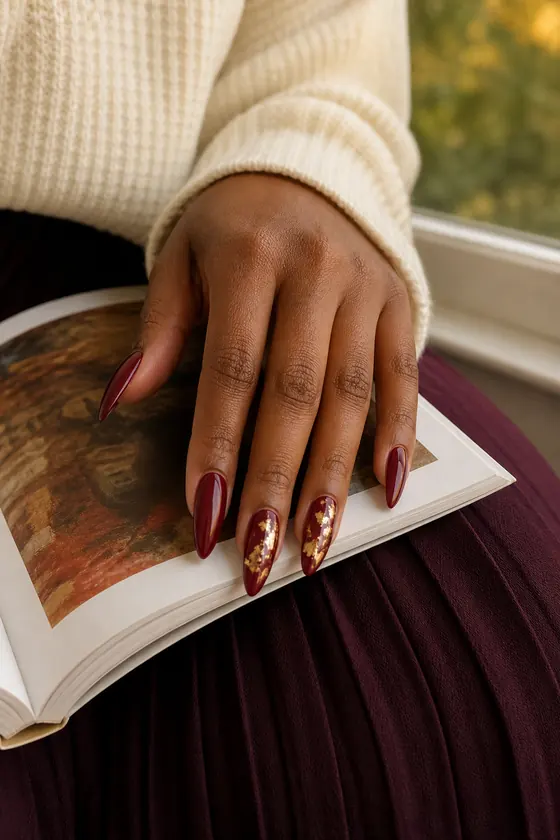 Long burgundy almond fall nails with gold foil on an open art book