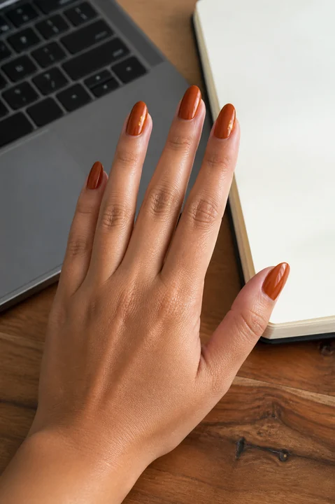 Glossy rust orange almond nails at an office desk