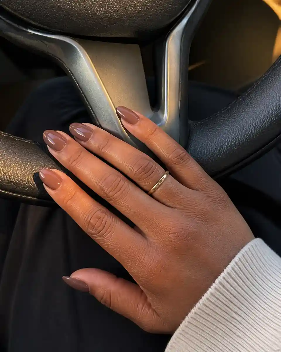 Glossy taupe nude almond nails resting naturally on a car steering wheel.