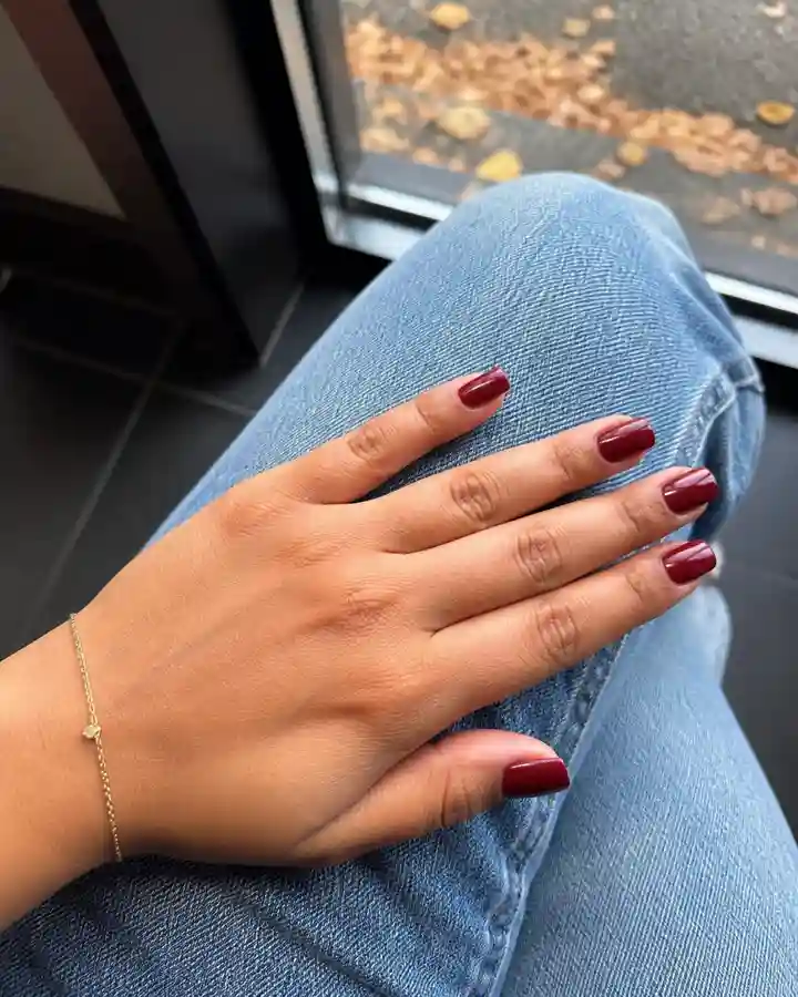 Short burgundy nails resting naturally on denim beside a cafe window.