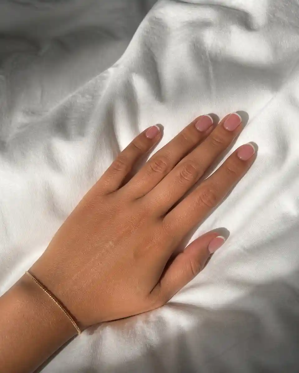 Precise thin French tip nails resting on white bed linen in morning light.