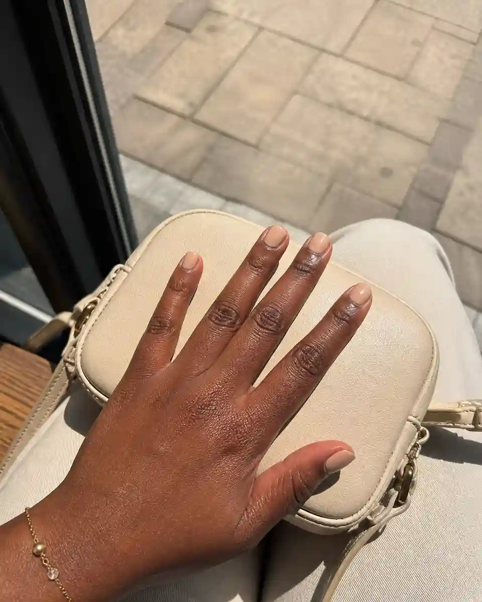 Short simple elegant nude nails on a Black woman beside a cafe window.