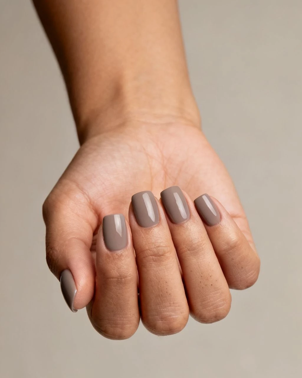 Short taupe-grey elegant manicure on a hand against a plain neutral studio background.
