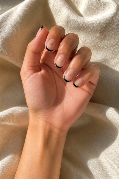 Black French tip nails with tiny gold stars on a light olive skin tone hand in an open palm angle with visible wrist and short partial forearm, natural daylight
