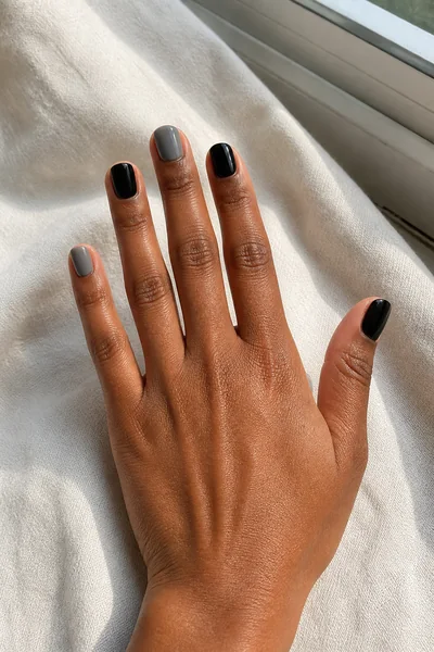 Short black and smoky grey alternating nails on a medium deep skin tone hand in a palm-down three-quarter pose with visible wrist and short partial forearm, natural daylight