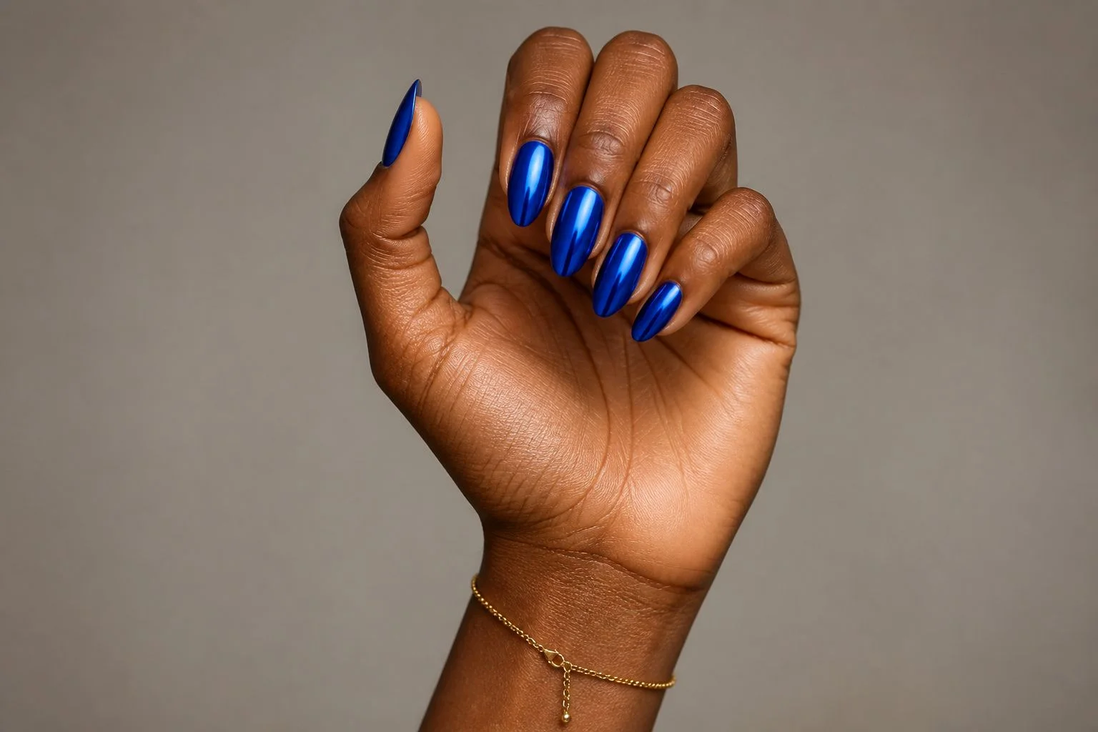 Curled hand showing a cobalt blue chrome mirror almond manicure with a delicate gold bracelet
