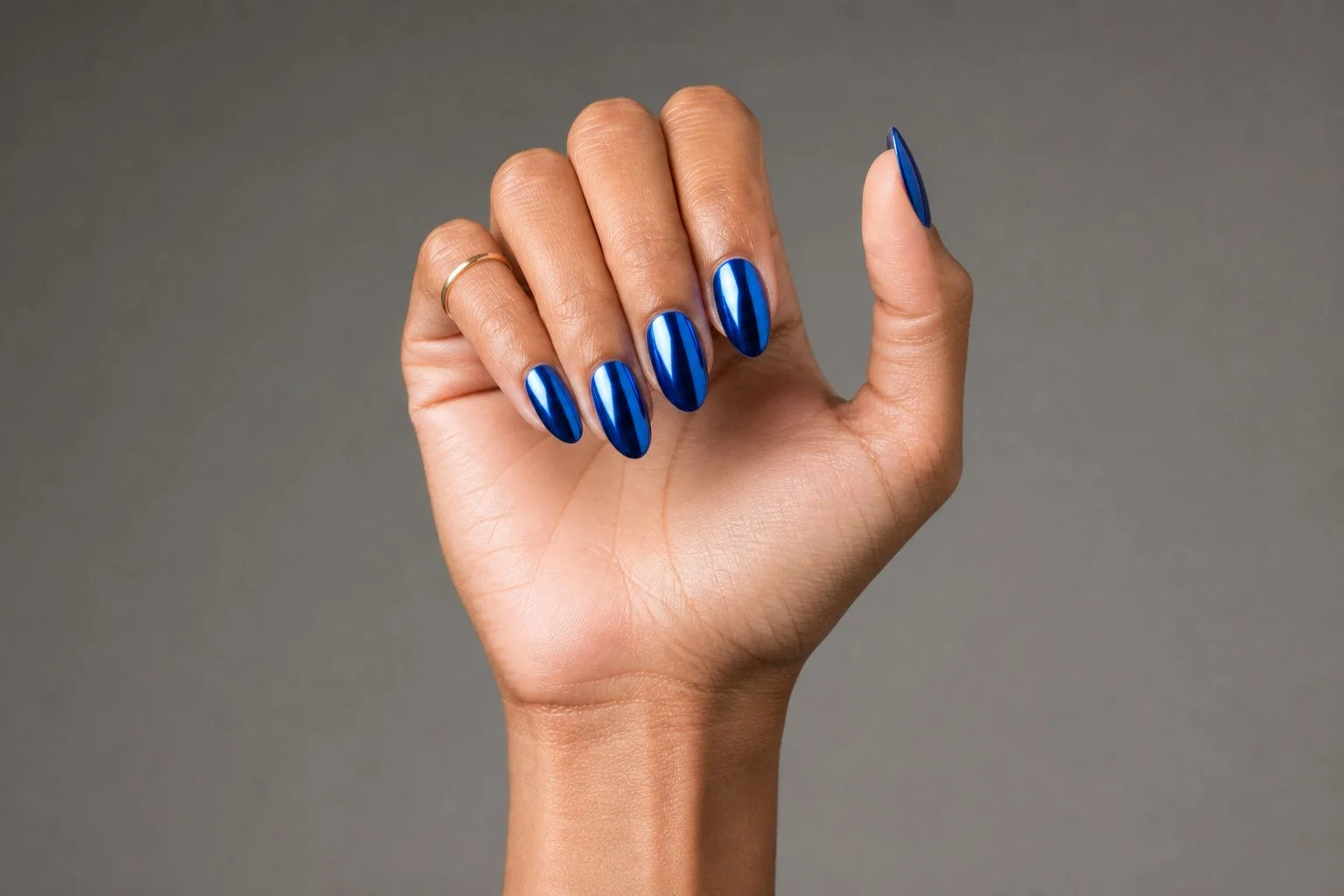 Curled hand showing a cobalt blue chrome mirror almond manicure with a delicate gold ring