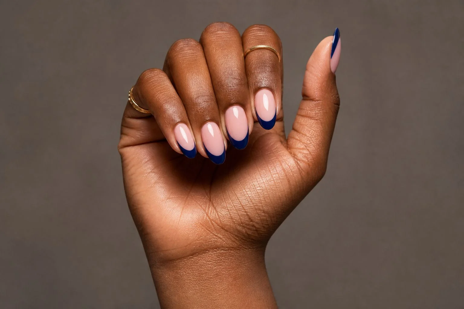 Curled hand showing a deep midnight blue almond French manicure with two thin gold rings