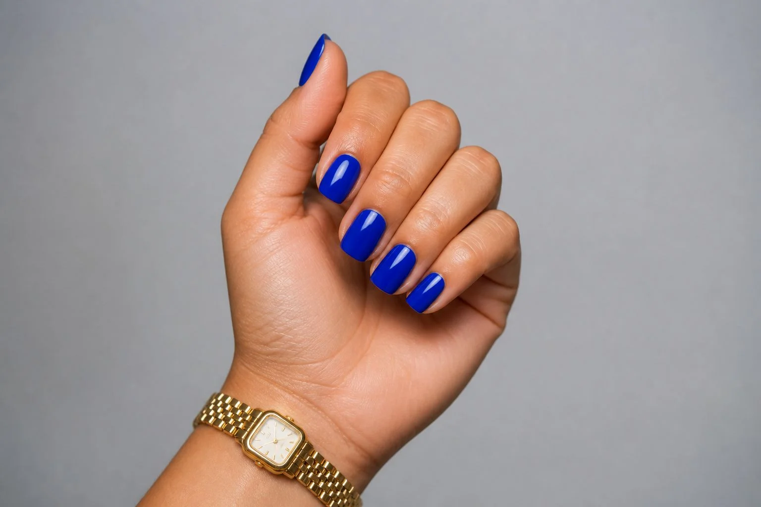 Curled hand showing a glossy royal cobalt blue square manicure with a slim gold watch
