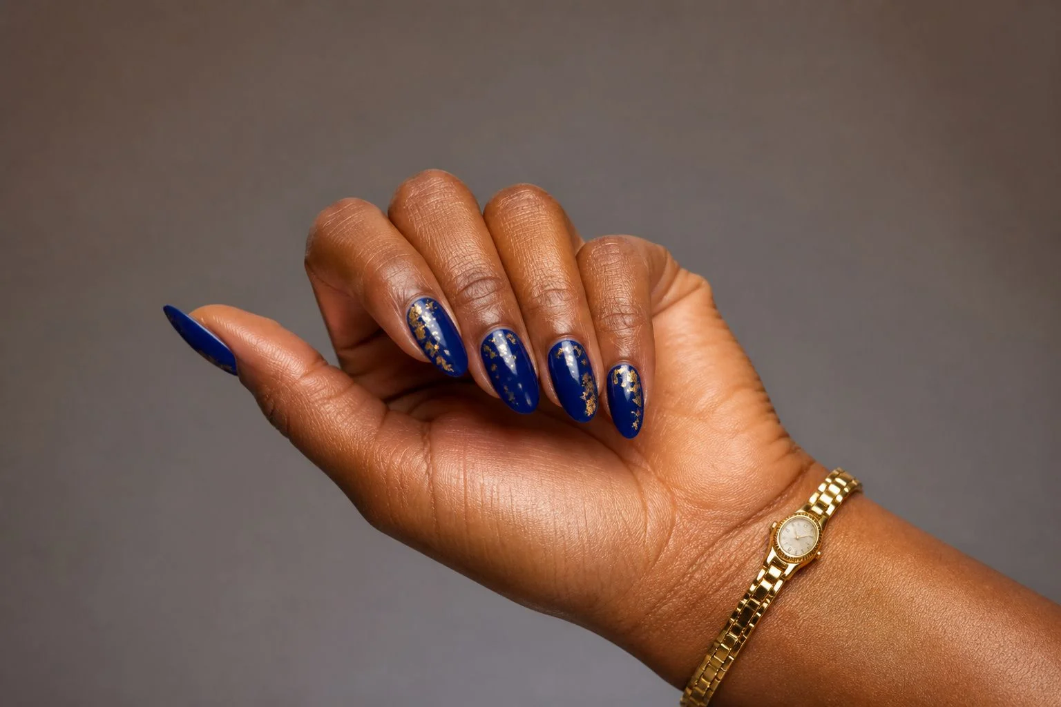Curled hand showing a glossy sapphire blue almond manicure with gold foil flake accents and a slim gold watch