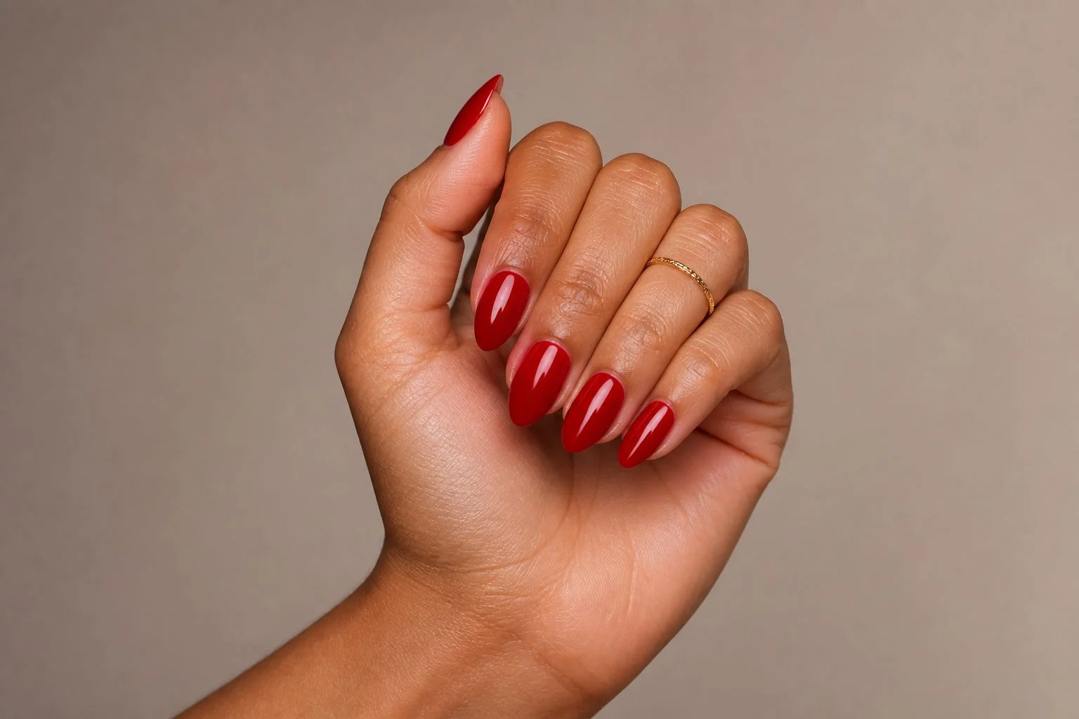 Curled hand showing a classic glossy red almond manicure with a delicate gold ring