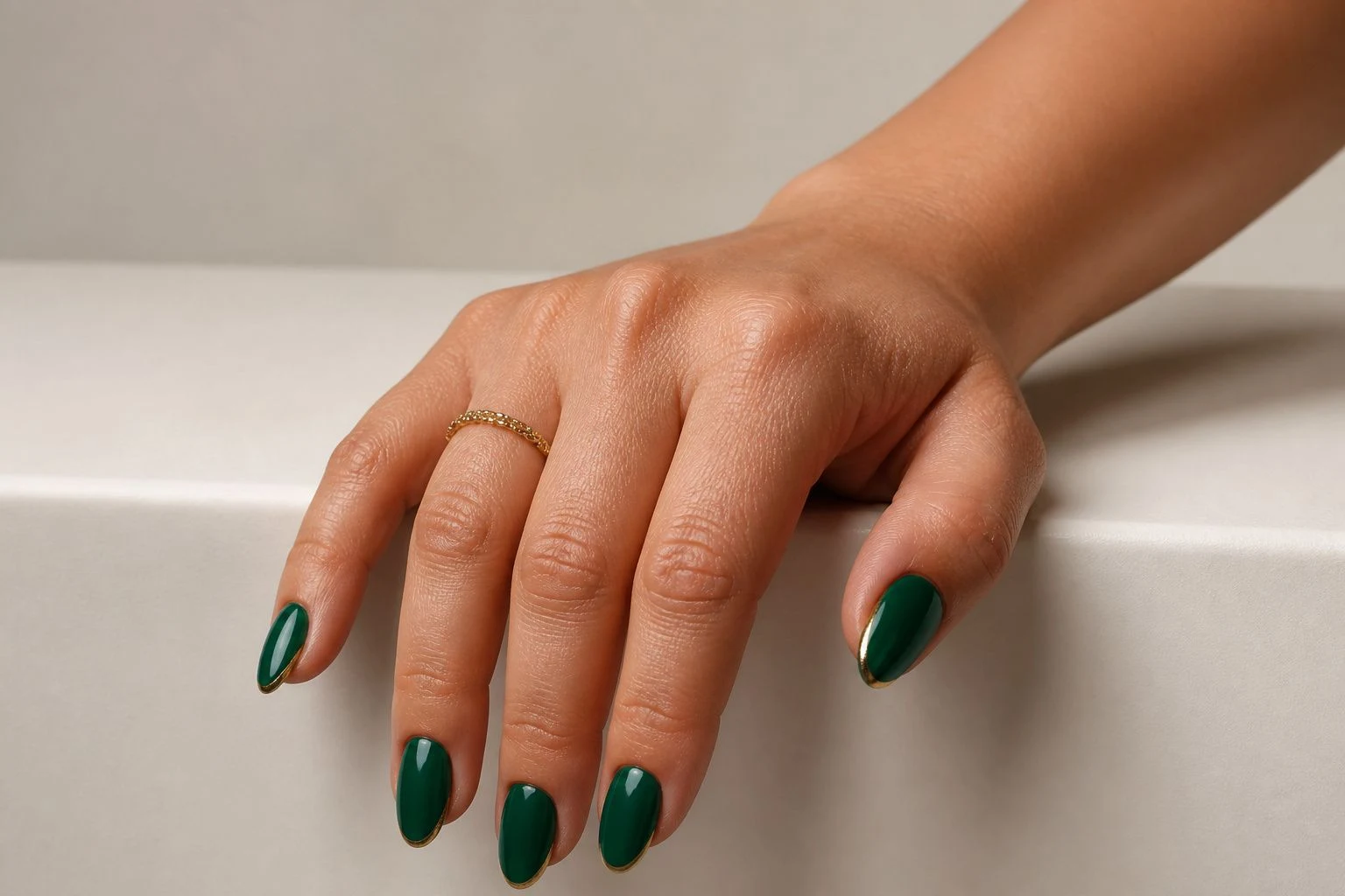 Emerald and gold almond French Christmas nails resting on a surface edge with a delicate gold ring