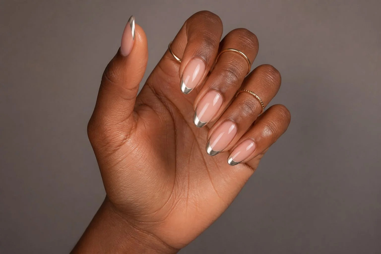 Curled hand showing a glossy silver chrome micro French almond manicure with two thin gold rings