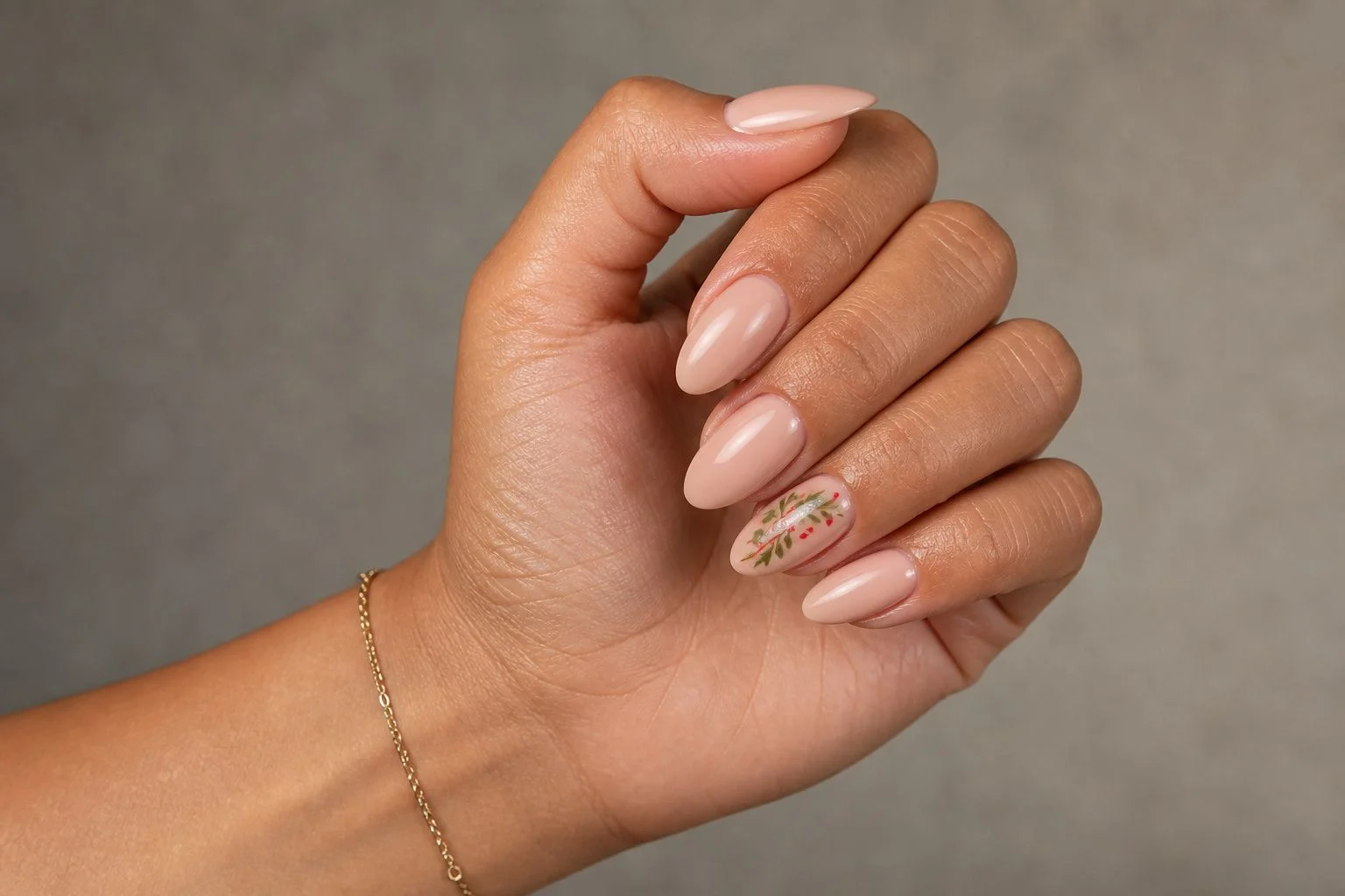 Elegant loose fist tilt showing a glossy nude almond manicure with one accent nail featuring refined red and green holiday art and a gold bracelet