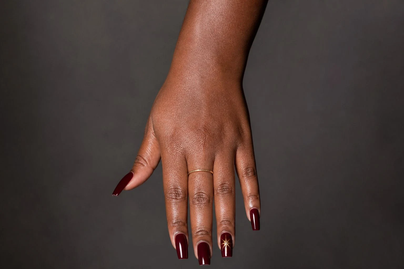 Long burgundy square Christmas nails with a star accent, wrist forward, with delicate gold rings