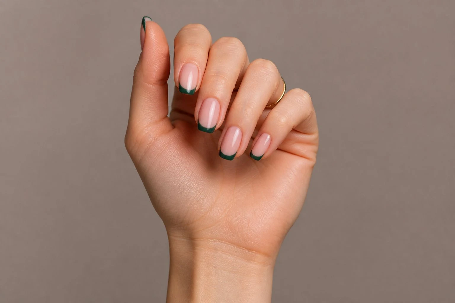 Green square French Christmas nails in a curled hand pose with a delicate gold ring