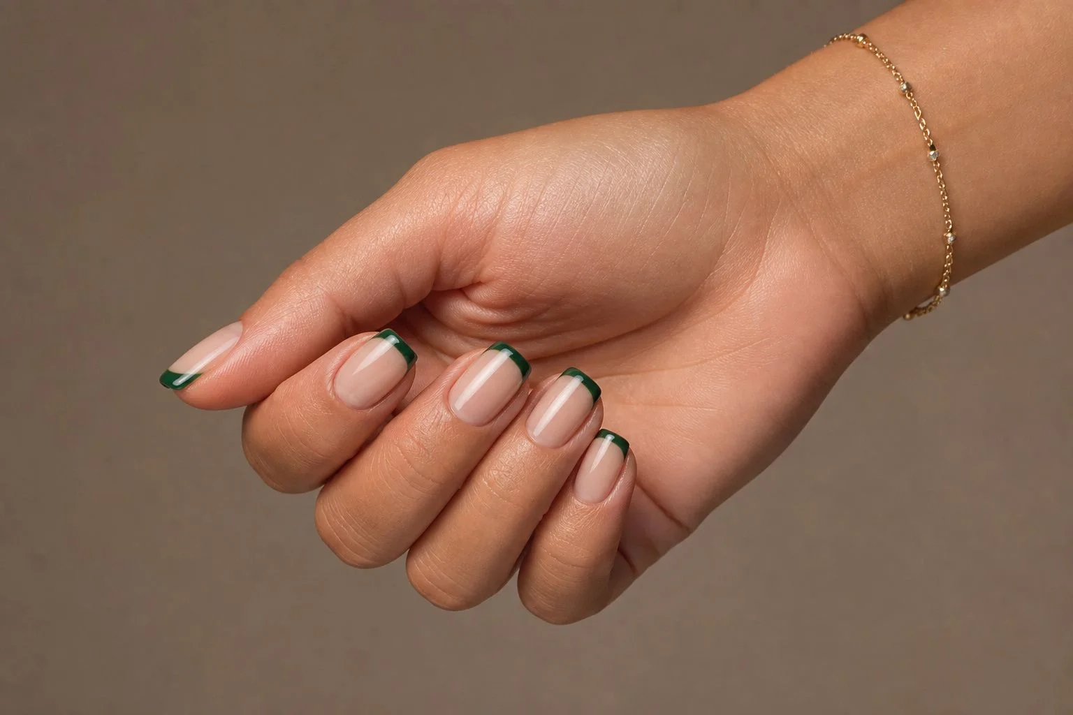 Emerald green square French Christmas nails with a delicate gold bracelet, relaxed lap pose