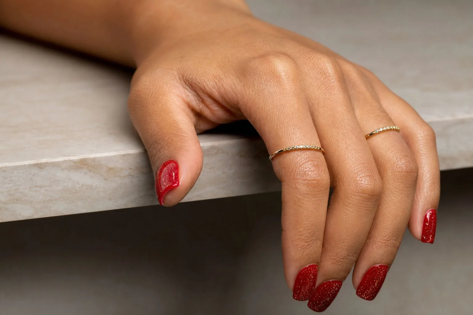 Red square Christmas nails with gold glitter accents and two thin gold rings, table edge pose
