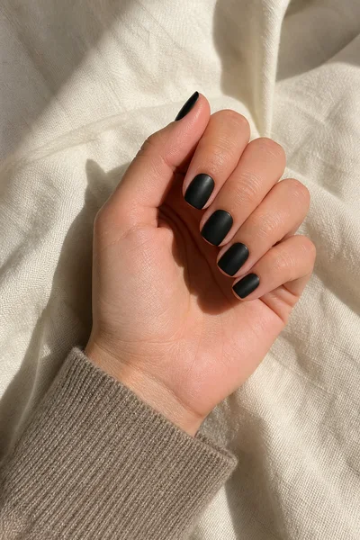 Black satin rounded square nails on a light olive skin tone hand in a compact softly curled pose, muted taupe knit cuff, visible wrist and short partial forearm, natural daylight