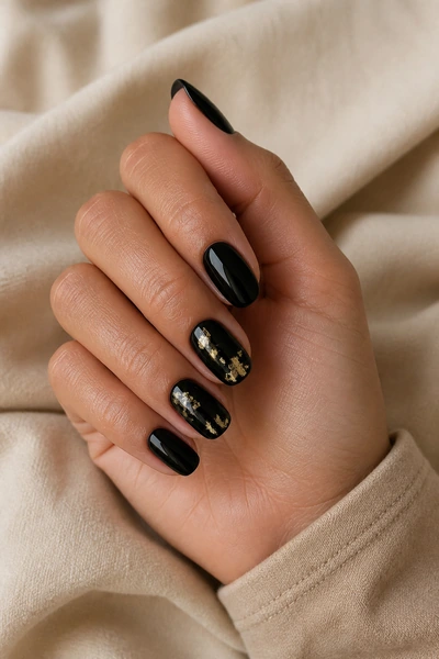 Black nails with small gold foil accents on a medium olive skin tone hand in a softly curled pose with visible wrist and short partial forearm, natural daylight