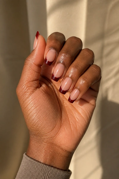 Deep red French tip duck nails on a rich dark brown skin tone hand in a gently curled pose with visible wrist and short partial forearm, natural daylight