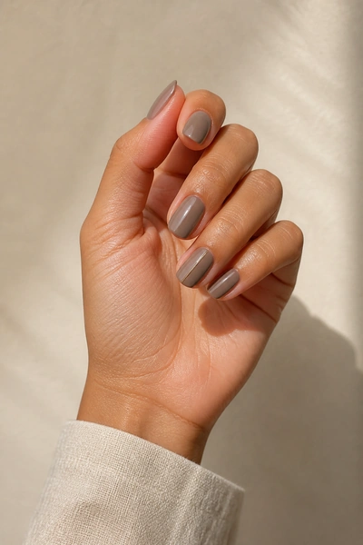 Short taupe duck nails with a fine gold accent on a tan skin tone hand in a gentle pinch pose with visible wrist and short partial forearm, natural daylight