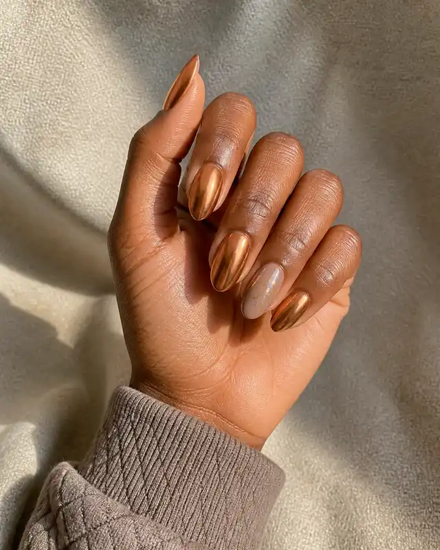 Burnished copper chrome medium almond nails with one smoky nude and tiny gold foil accent on a deep rich warm-brown skin tone hand in a softly curled pose with a taupe quilted cuff, visible wrist and short partial forearm, natural daylight