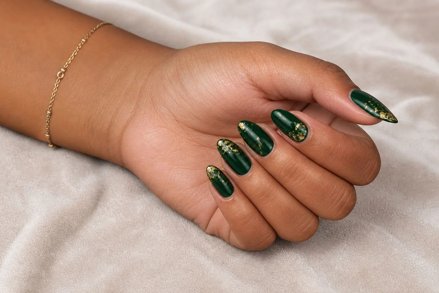 Soft closed fist showing a glossy emerald green almond manicure with gold foil flake accents and a gold bracelet