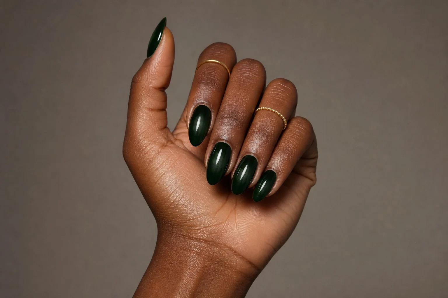 Overlapping curled fingers showing a glossy forest green almond manicure with a deep ombre fade and two thin gold rings