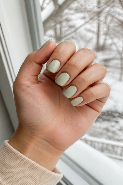 Soft sage green rounded square nails on a light olive skin tone hand in an open palm angle with visible wrist and short partial forearm, natural daylight