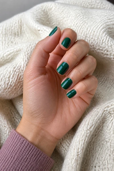 Short glossy emerald square nails on a light brown skin tone hand in a gentle pinch pose with visible wrist and short partial forearm, natural daylight