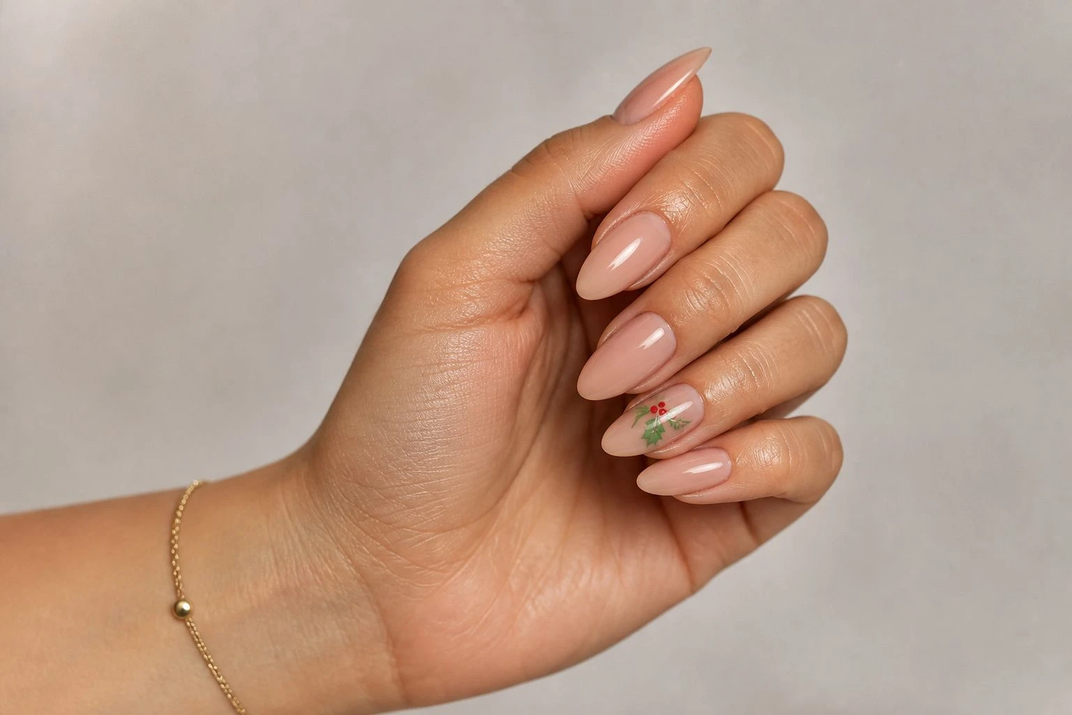 Glossy nude almond nails with a holly leaves and berries accent nail and a gold bracelet, Christmas nail idea
