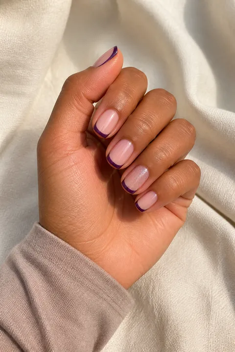 Deep violet French tips with one silver shimmer accent on short squoval nails, a medium warm-brown skin tone hand in a softly curled pose with a taupe sleeve, visible wrist and short partial forearm, natural daylight