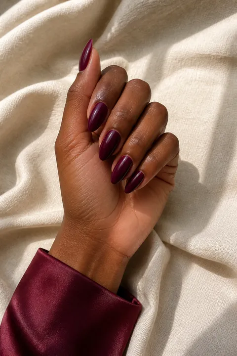 Glossy burgundy plum medium almond nails on a deep warm-brown skin tone hand in a graceful softly curled pose with a burgundy satin cuff, visible wrist and short partial forearm, natural daylight