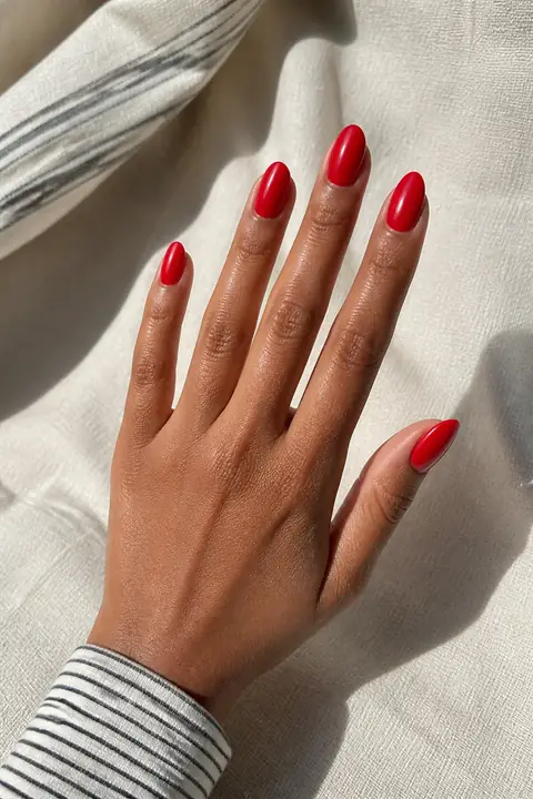 Glossy red medium-short almond nails on a medium warm-brown hand in an open diagonal pose with a narrow-stripe cuff