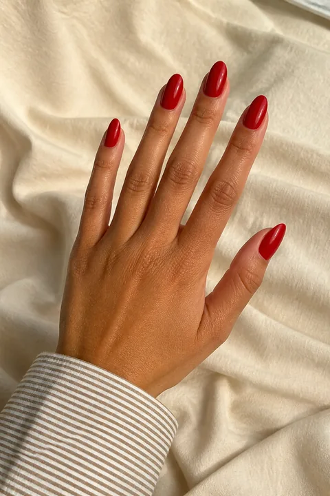 Bright glossy cherry-red medium almond nails on a tan golden hand in an open diagonal pose with a narrow-stripe cuff