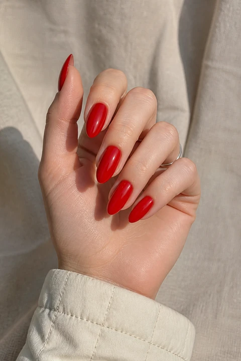 Glossy deep red almond nails on a light beige hand in a compact curled pose with a cream quilted cuff and tiny silver ring