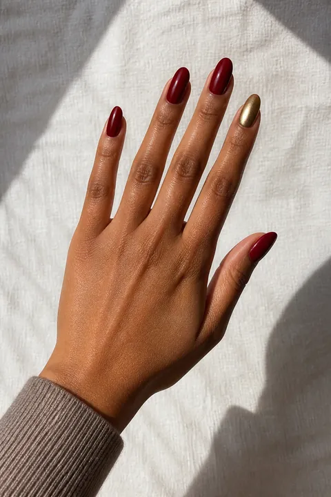 Deep red glossy medium-long almond nails with one metallic gold ring-finger accent on a medium-deep brown hand in an open diagonal pose with a taupe knit cuff