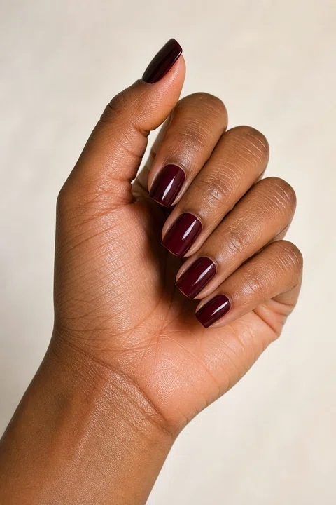 Deep neutral brown skin hand in a curved radial pose with five short square glossy oxblood burgundy acrylic nails