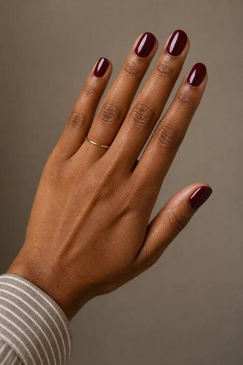 Medium-deep warm brown skin hand in an open diagonal pose with five short rounded mirror-gloss burgundy nails, a narrow-stripe cuff, and one tiny plain gold band