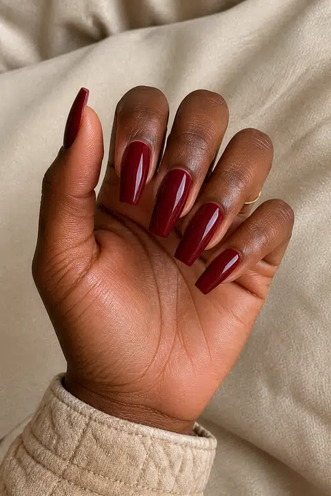 Rich dark brown skin hand in a layered fan pose with five medium tapered coffin nails painted glossy deep burgundy wine red, a beige quilted cuff, and one tiny plain gold ring