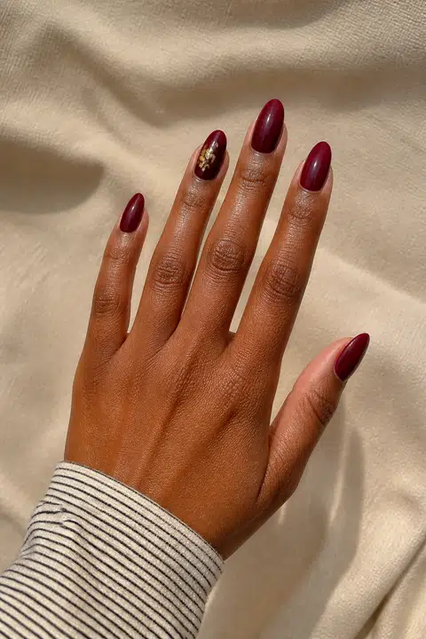 Medium-deep warm brown skin hand in an open diagonal pose with five medium almond glossy deep burgundy nails and one delicate gold foil accent on the ring nail, wearing a neutral narrow-stripe cuff