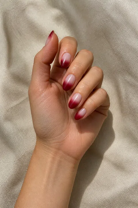 Light olive skin hand in a controlled curled pose with five short almond nails in a soft glossy blush nude to deep burgundy ombre against beige textile