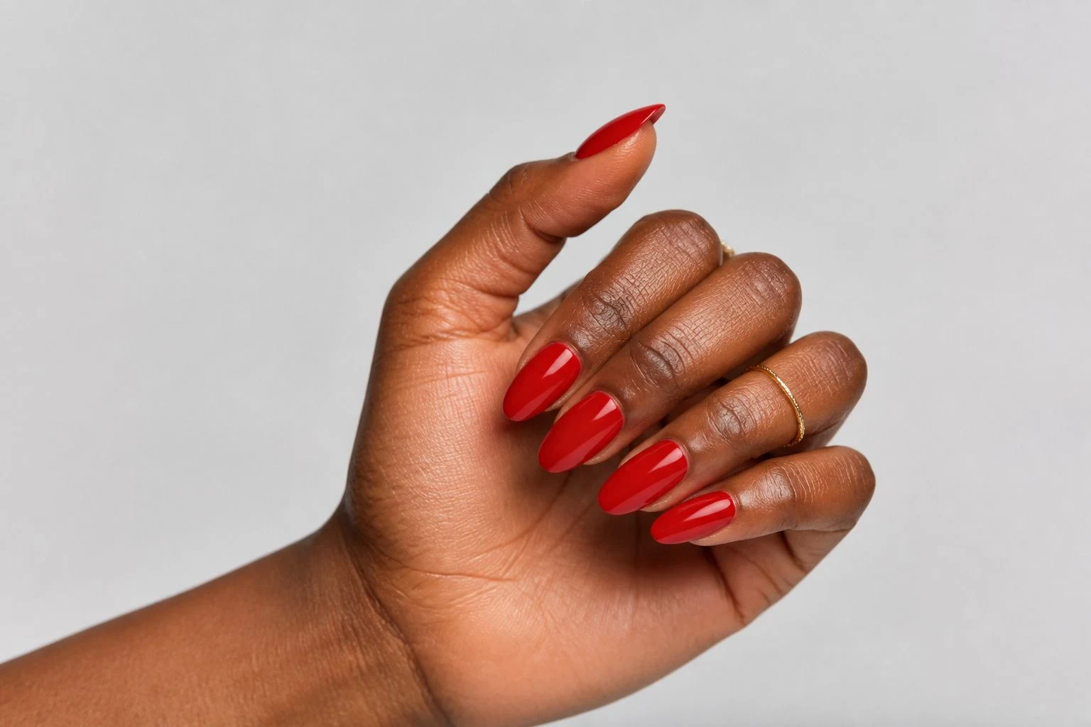 Candy-apple red almond Christmas nails in a curled hand pose with a delicate gold ring