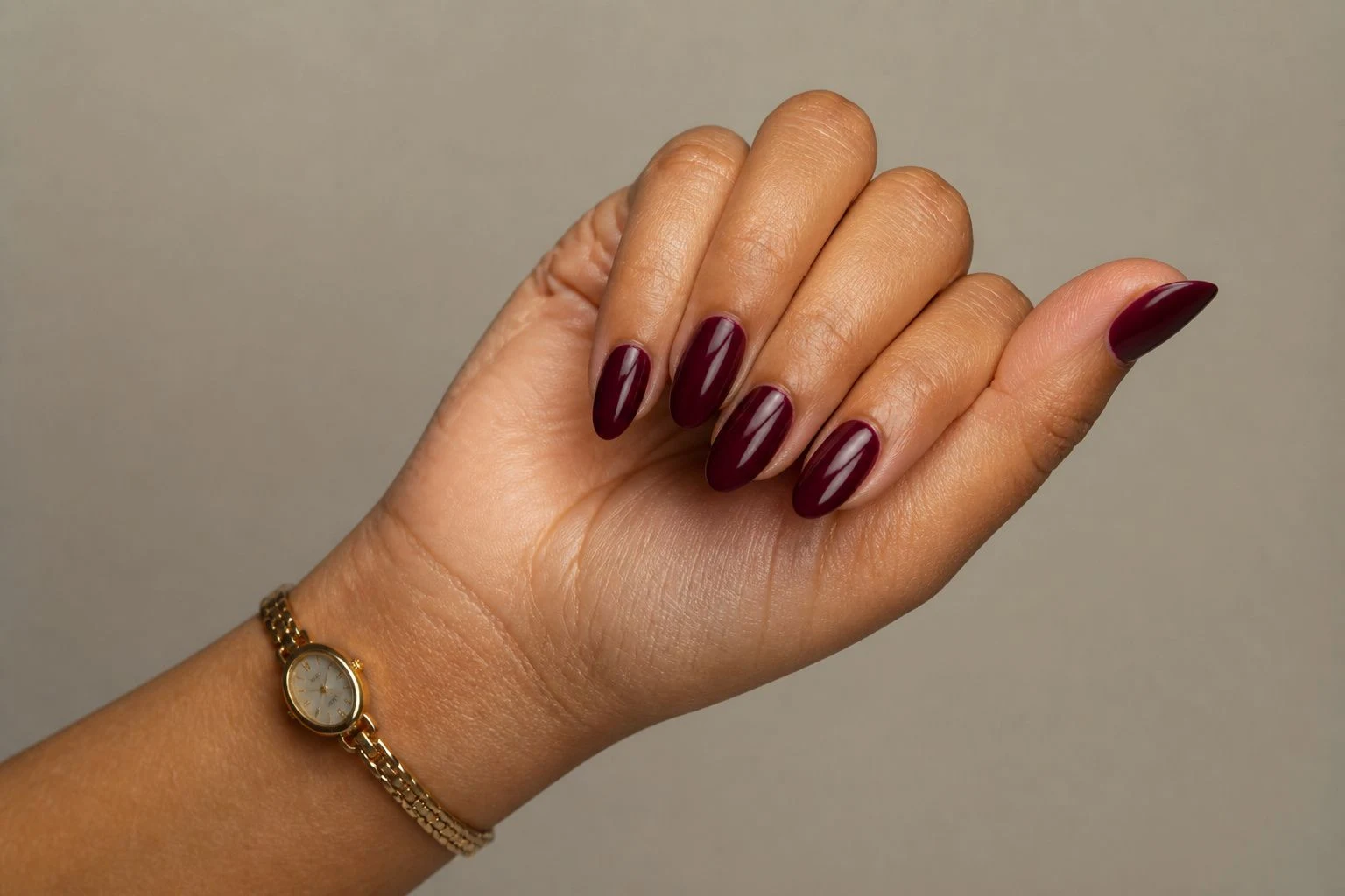 Deep wine almond Christmas nails in a curled hand pose with a delicate gold watch