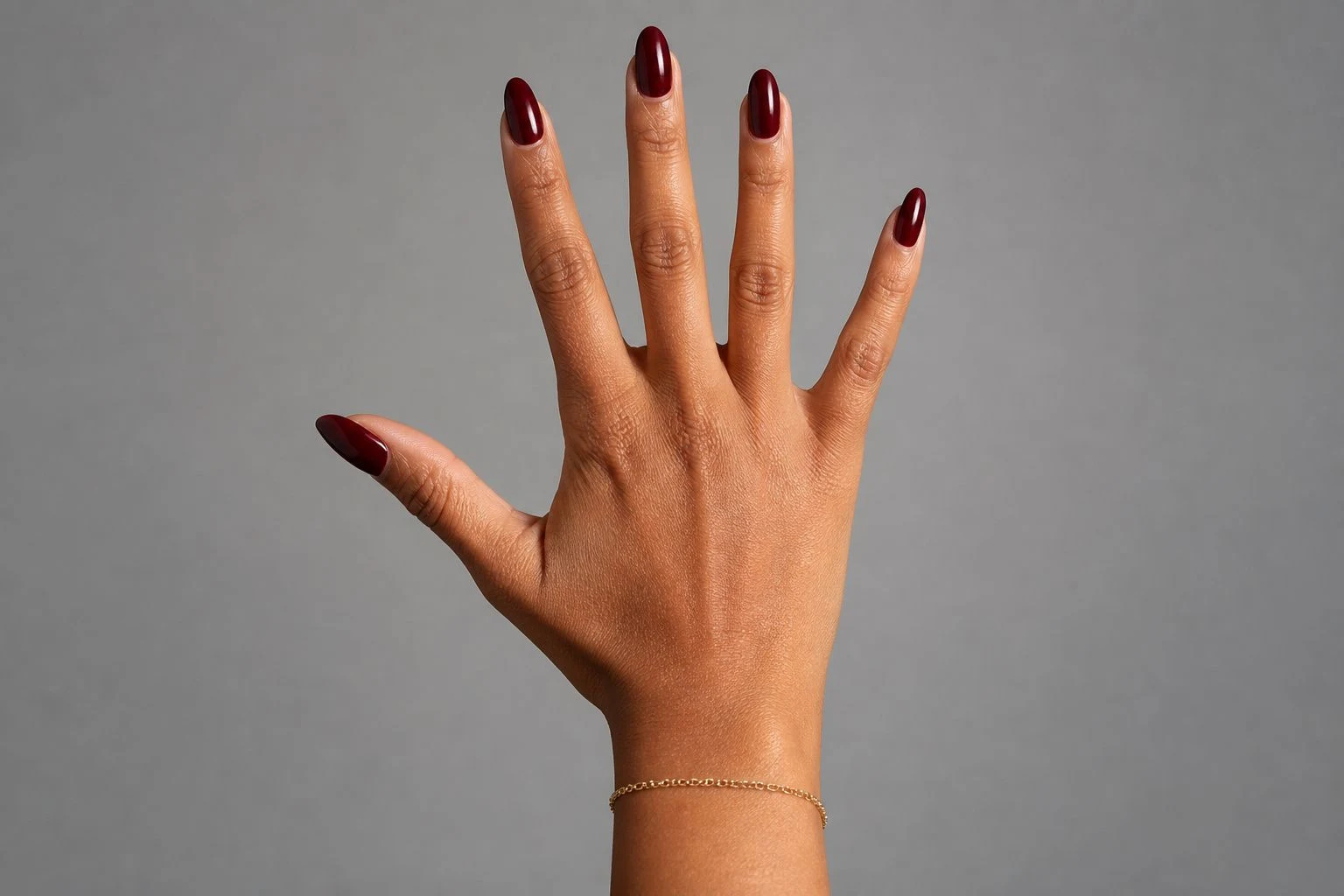 Deep wine red almond Christmas nails in an open palm pose with a delicate gold bracelet