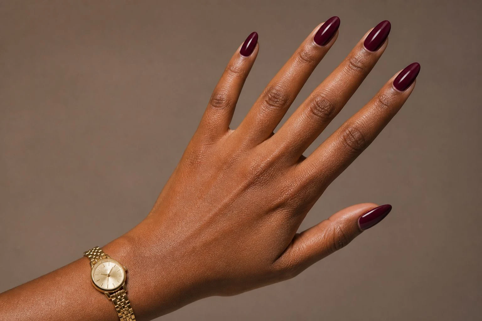 Deep wine almond Christmas nails in a diagonal hand pose with a delicate gold watch