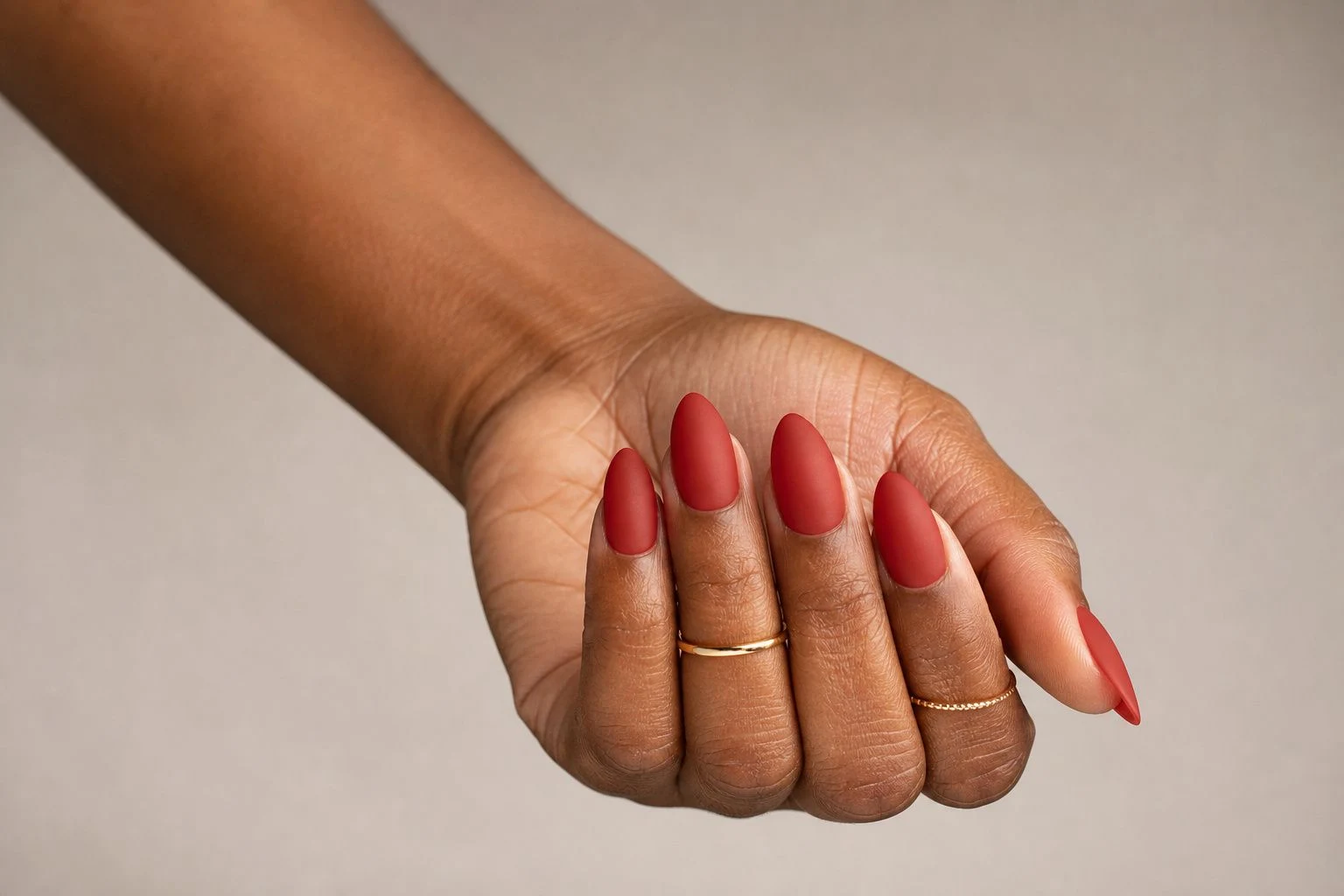 Matte red almond Christmas nails in a curled hand pose with delicate gold rings