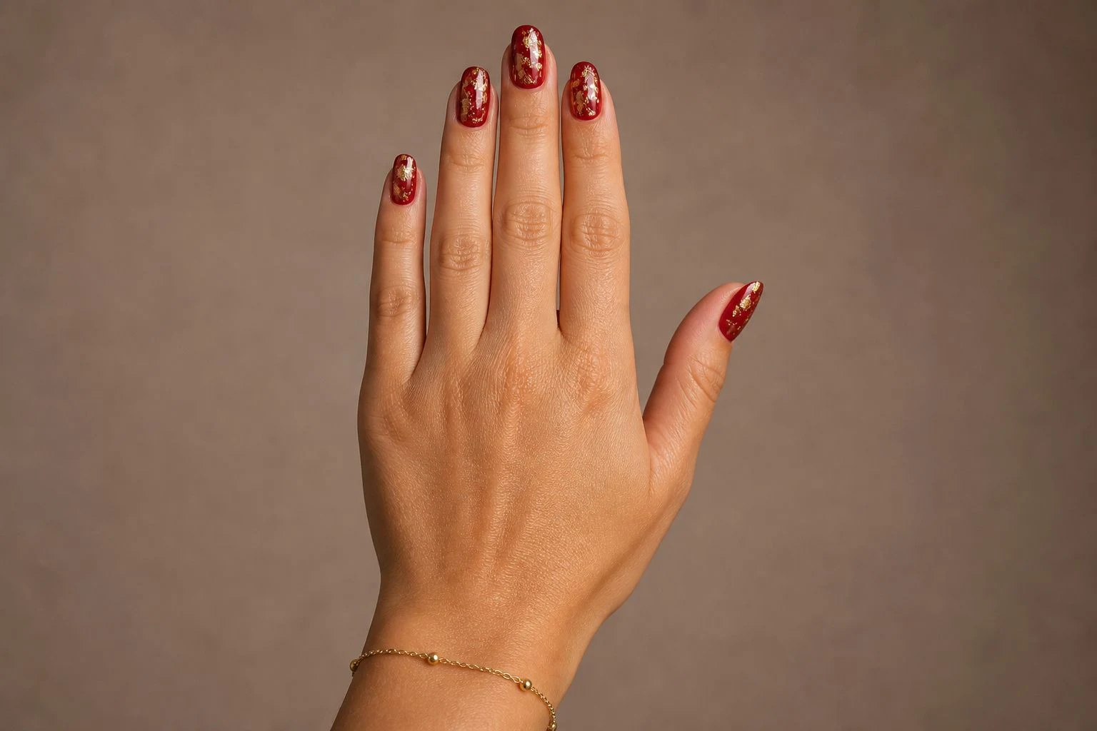 Red Christmas nails with refined gold foil in a back-of-hand pose with a delicate gold bracelet