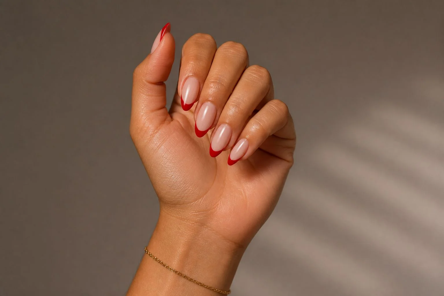 Curled hand showing a glossy nude almond manicure with a classic red French tip accent and delicate gold bracelet